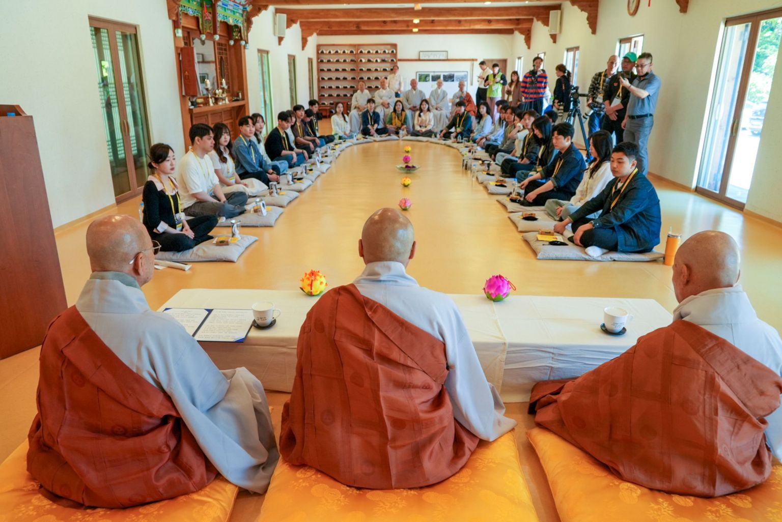 大批参加者坐在寺院大殿内的圆形坐垫上，围成半圆，前方有三名僧人。