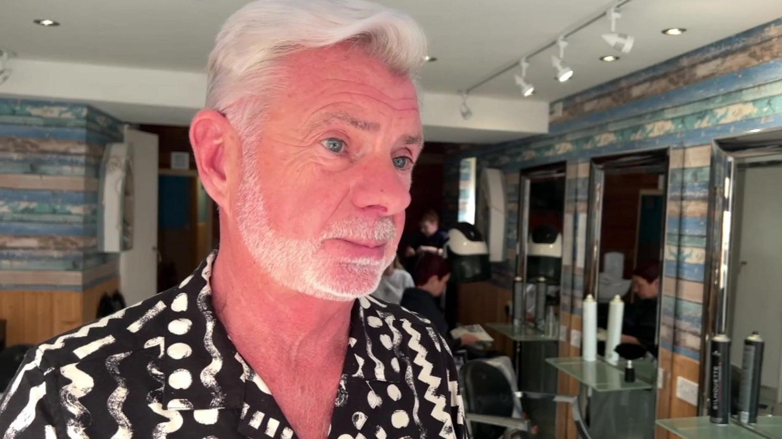 Michael Elliott-Jones standing in his salon. He has short white hair and a neatly-trimmed full beard and is wearing a black shirt with white zig-zag and dot patterns