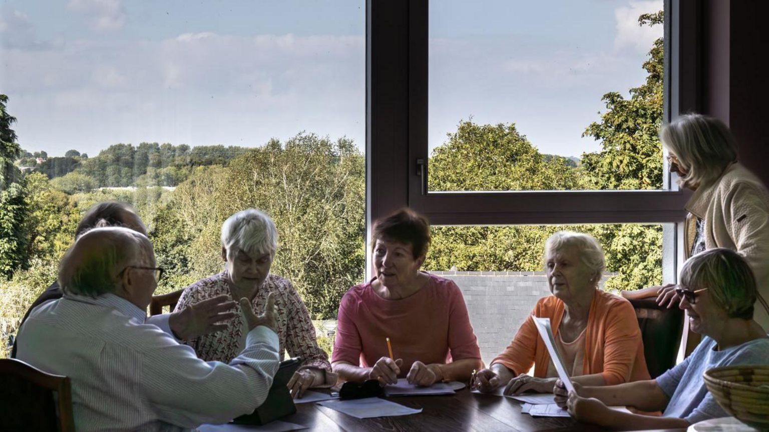 Six retirement village residents sit around a table in front of a big window. They have pens and paper in their hands. One woman stands behind one of the resident's chairs