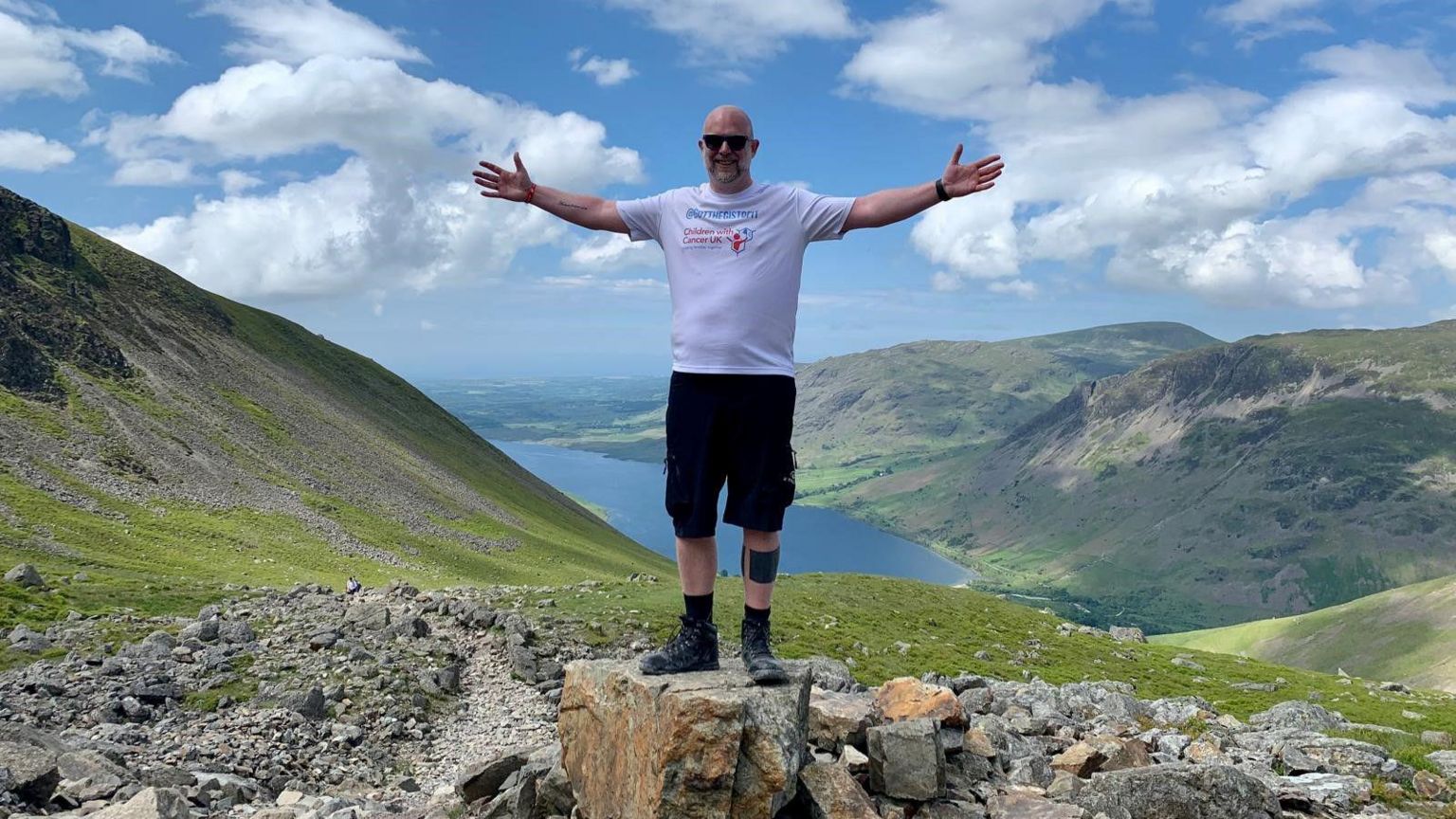 Chris Johnson standing at the top of a mountain with a lake below him. He has his hands in the air 