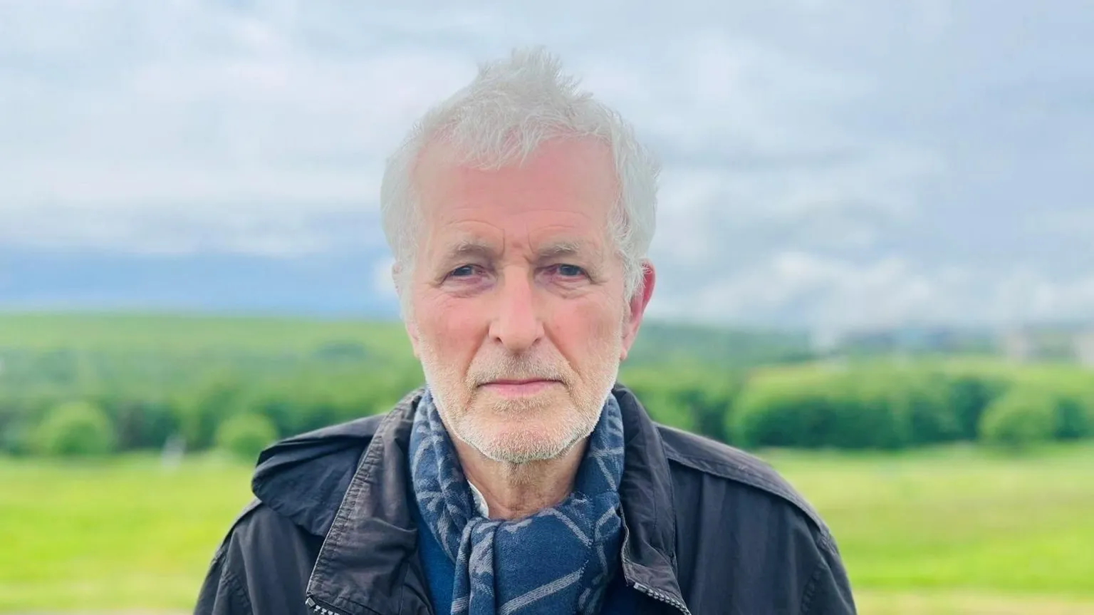Ian Baird, a man with grey hair and grey stubble, looking at the camera, he is wearing a dark jacket and a blue scarf, with a grassy landscape behind him.