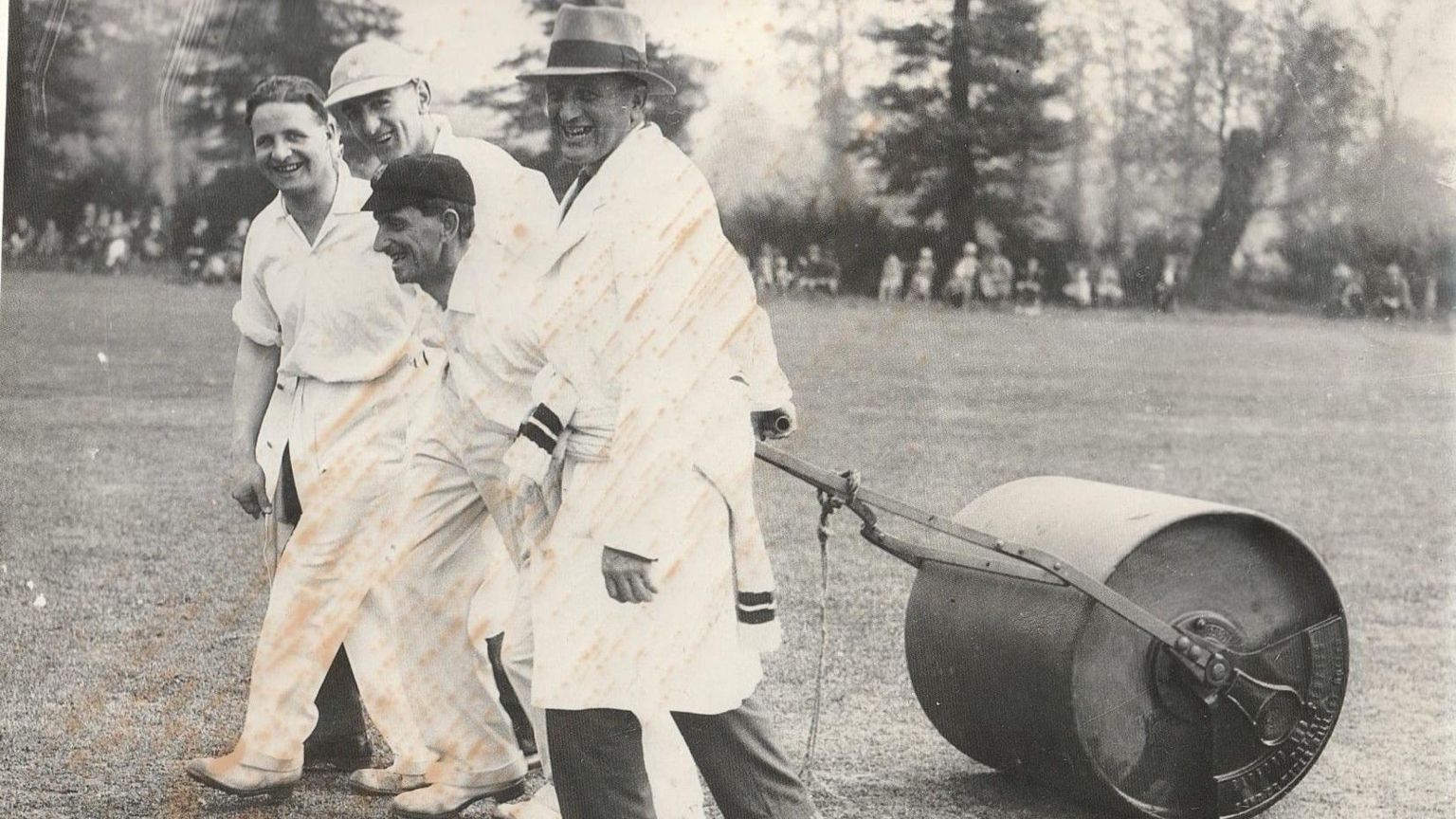 Four men pulling cricket pitch roller in black and white photo