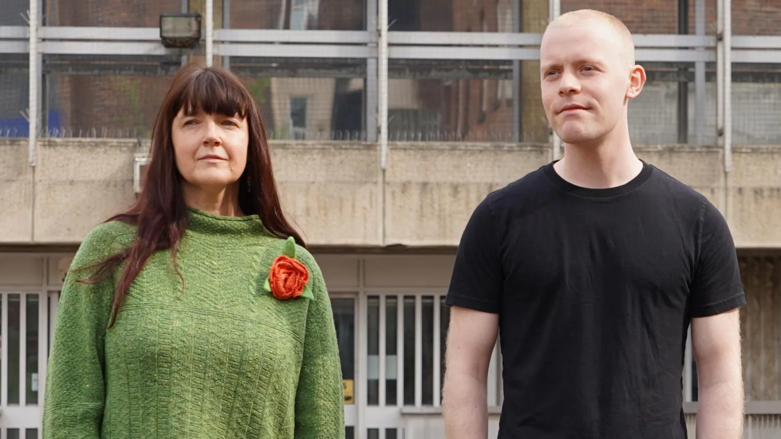 Two people stand outside a modern building with glass windows. On the left is a woman with dark hair and bangs wearing a green knitted sweater with an orange flower decoration. On the right is a bald man in a black t-shirt. Both are looking slightly upward.