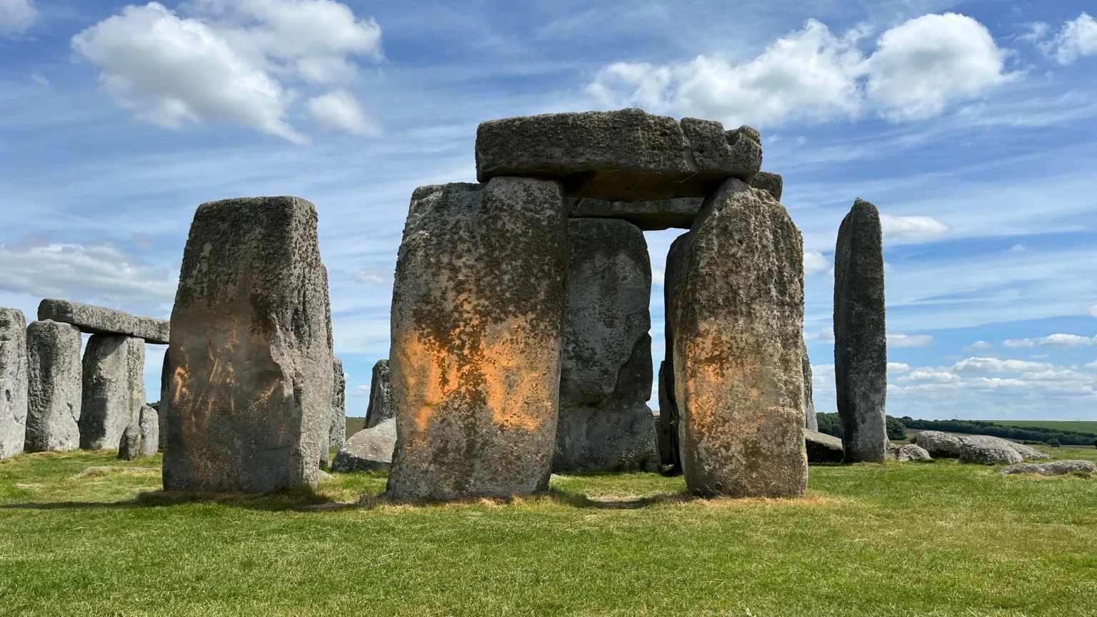 This image shows Stonehenge with bright orange powder paint splattered across several of the ancient stone trilithons. The prehistoric monument stands on green grass under a cloudy blue sky. The orange substance is clearly visible on multiple stones, contrasting starkly against the grey sarsen stones of this famous archaeological site.