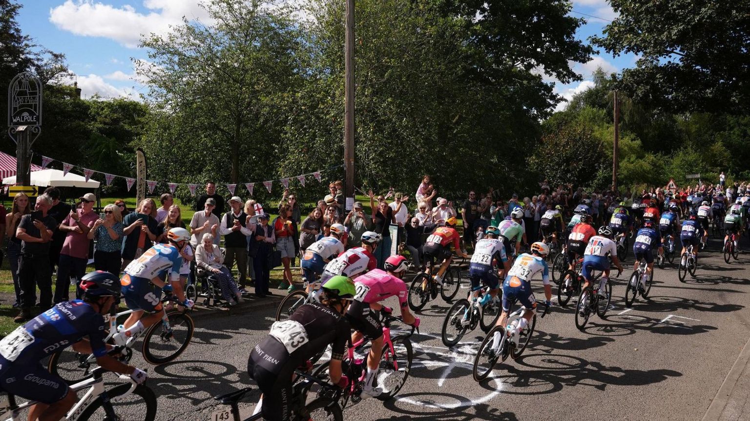 The view from behind of many cyclists in colourful outfits and helmets racing through Walpole Village, with the Walpole sign to the left and crowds lining the street