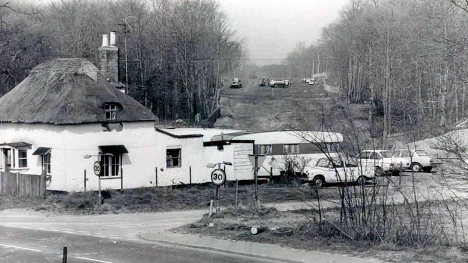 A black and white photo of a house with a caravan and cars parked to the back of it, with construction going on in the background. 