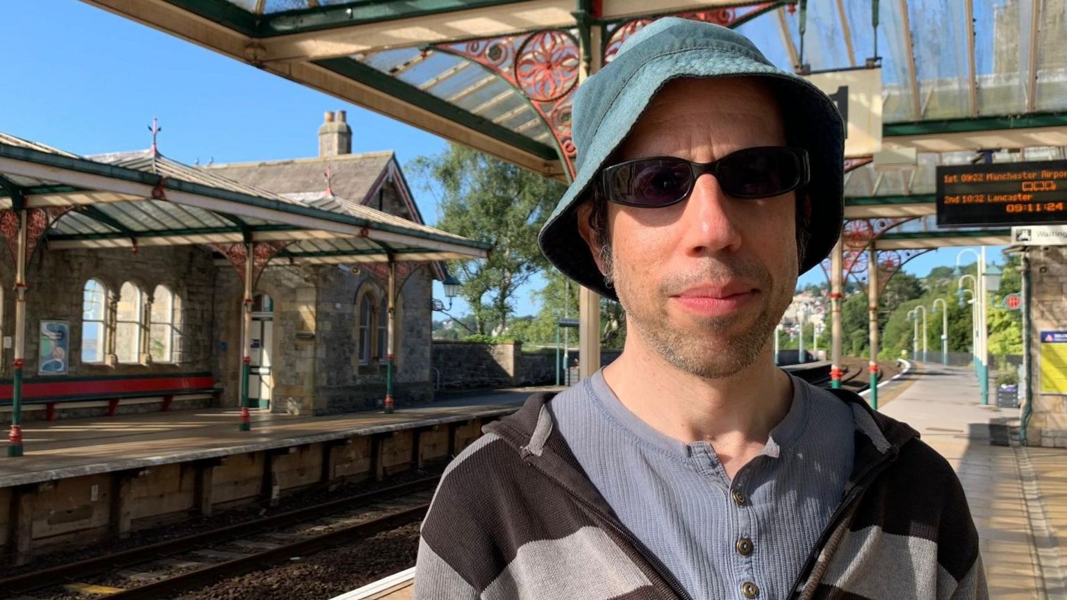 A man wearing sunglasses and a green bucket hat stands on a railway platform.