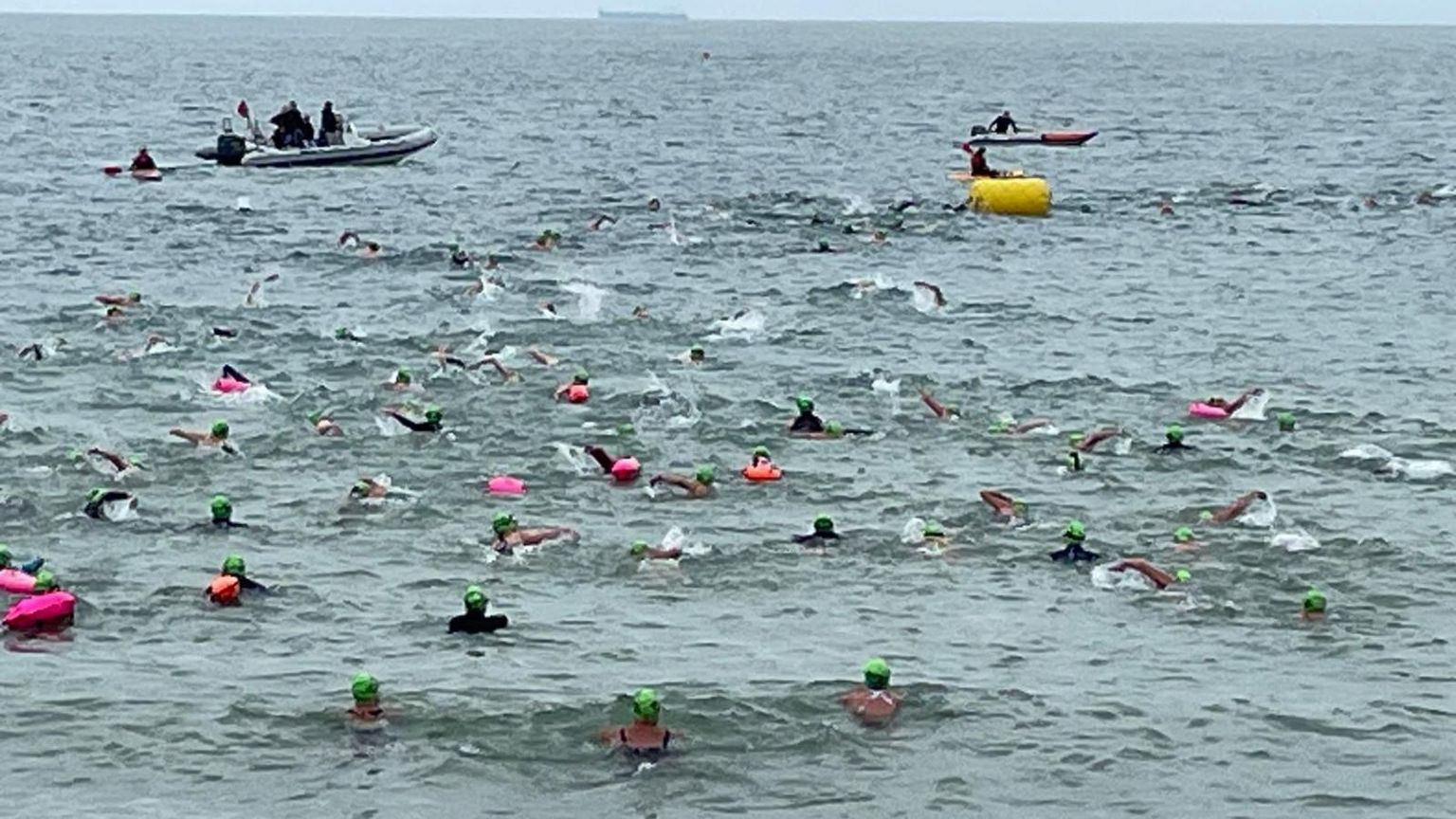 Swimmers in the water with various different coloured caps visible just above the water and support boats can be seen in the distance