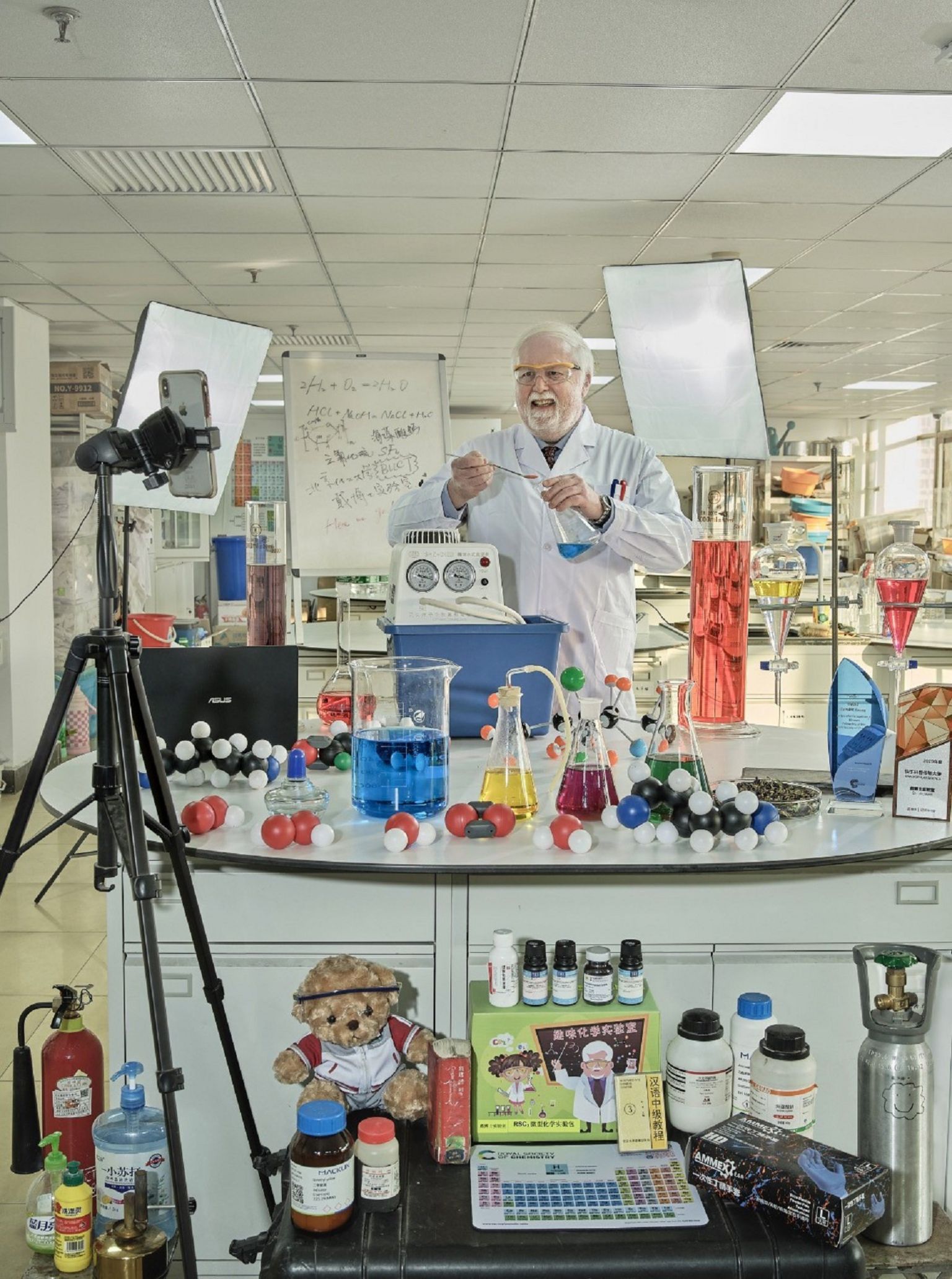 Профессор стоит перед столом в лаборатории, заставленной мензурками, цилиндрами, оборудованием для титрования и другим лабораторным оборудованием, наполненным разноцветными жидкостями