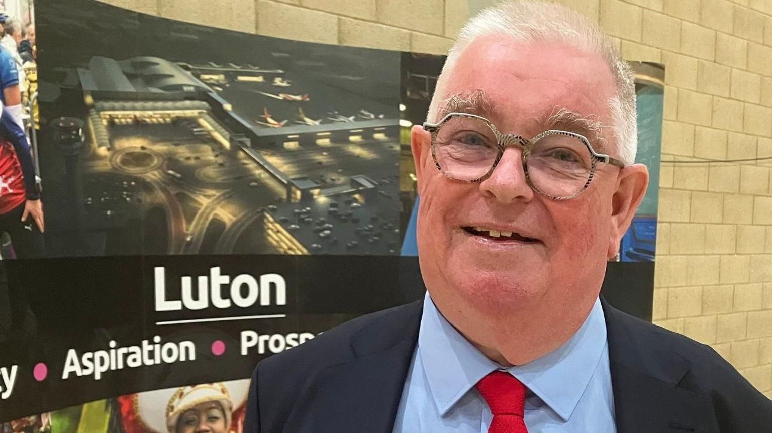 John Tizard smiling in front of a sign that says "Luton"