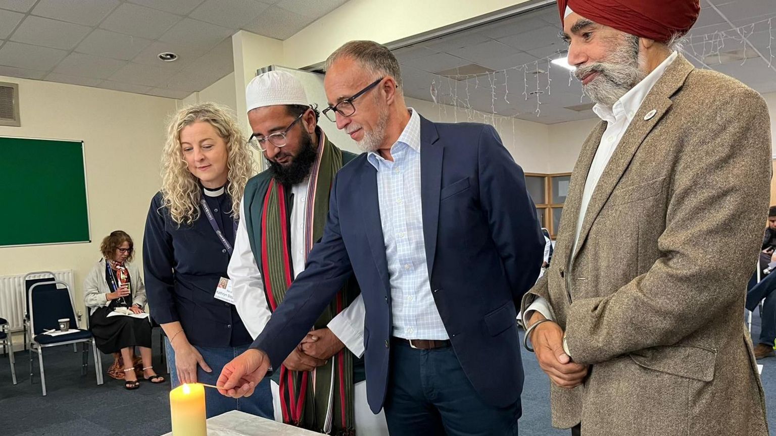 A woman and three men of different faiths gather round a candle as one of the men lights it