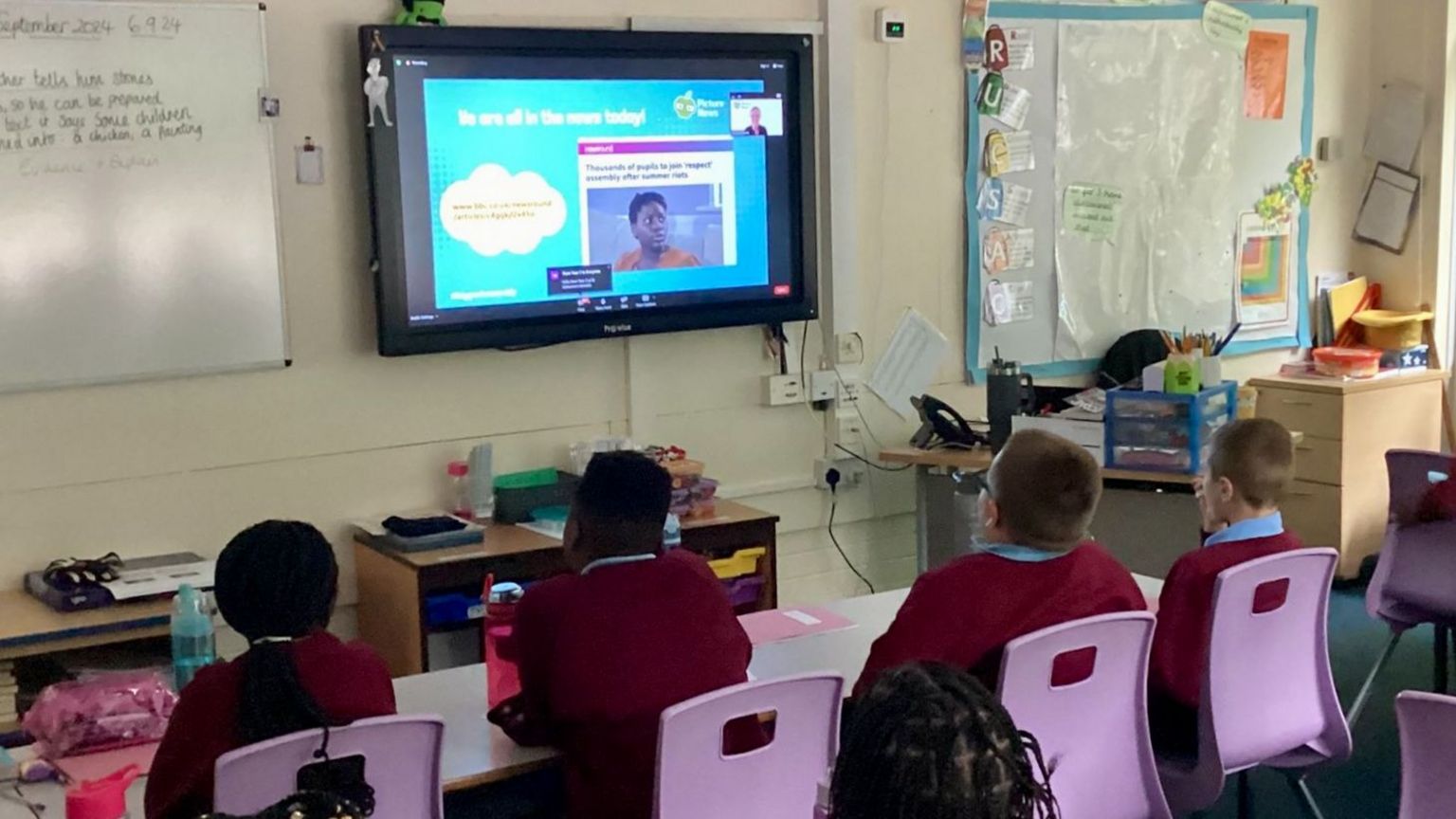 Primary-aged children in red jumpers  sit at rows of desks with their backs to the camera. The Big Back to School Assembly is being shown on the interactive whiteboard on the wall.