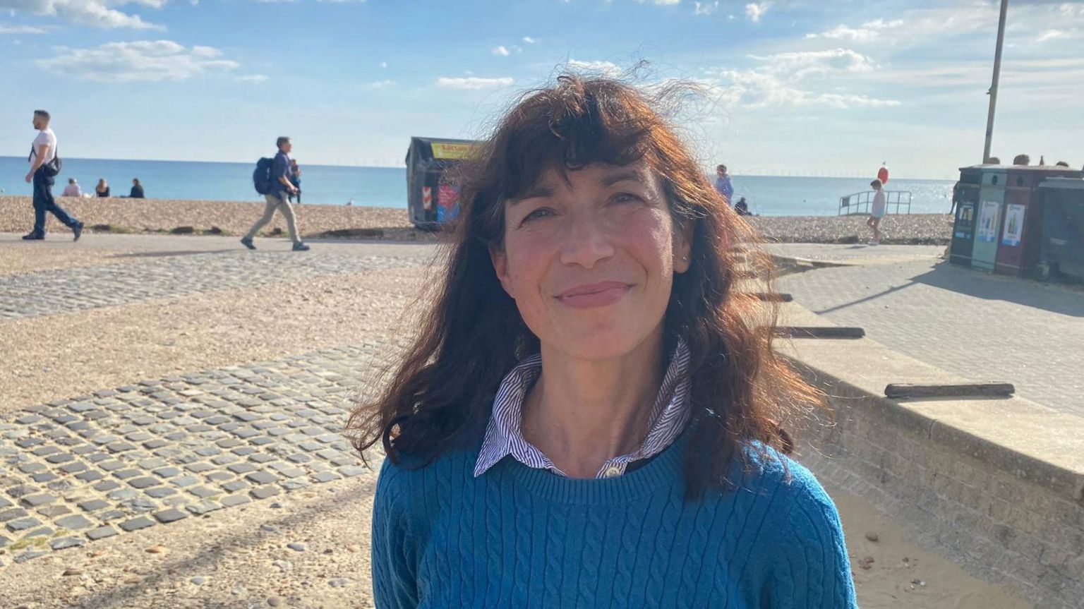 She has brunette shoulder length hair with a fringe. She's wearing a collared, blue and white pinstripe shirt underneath a light blue cable neck jumper.