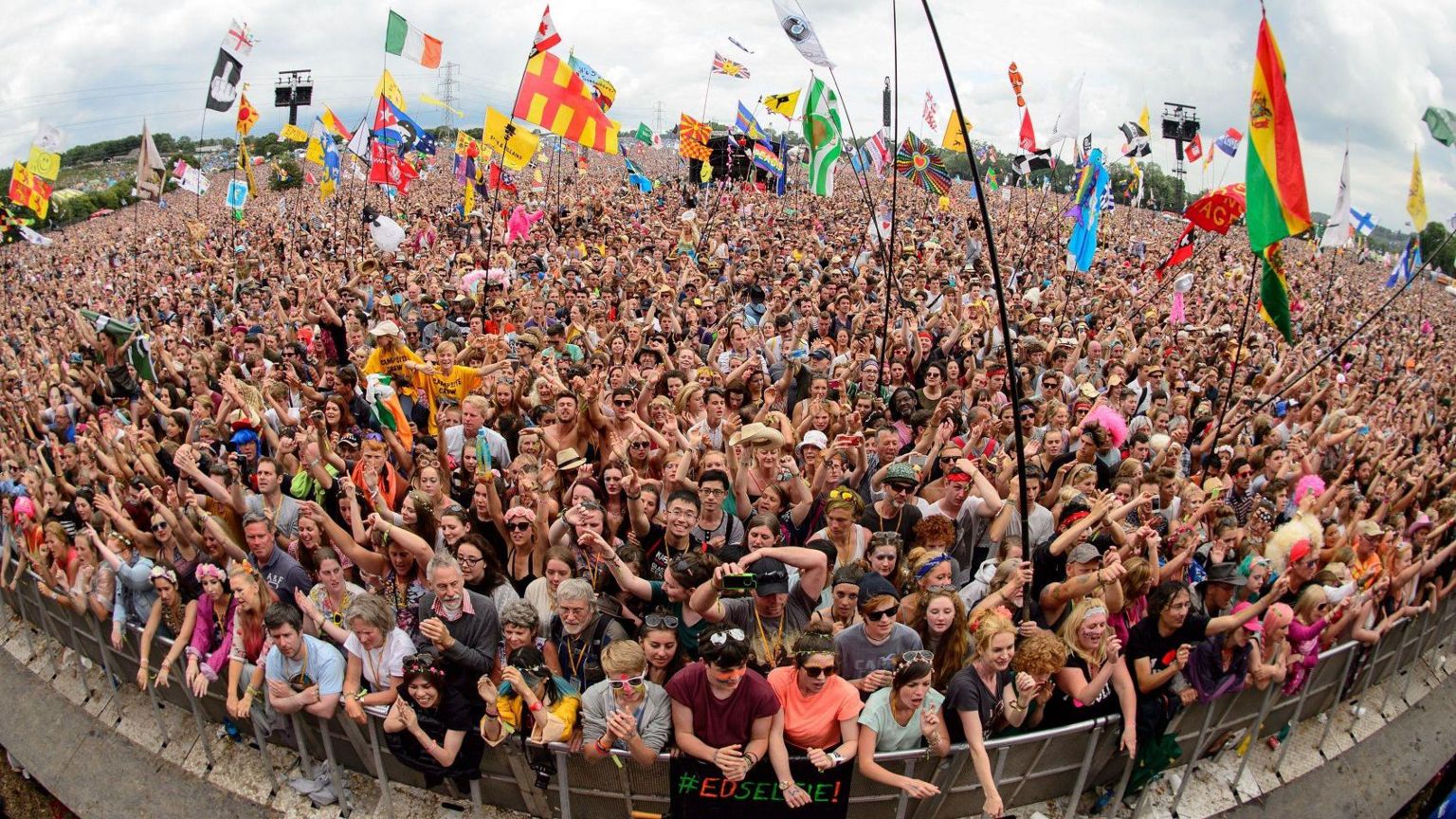 A large crowd packed in, in front of the Pyramid Stage at Glastonbury. People are leaning on the crash barrier and also waving flags