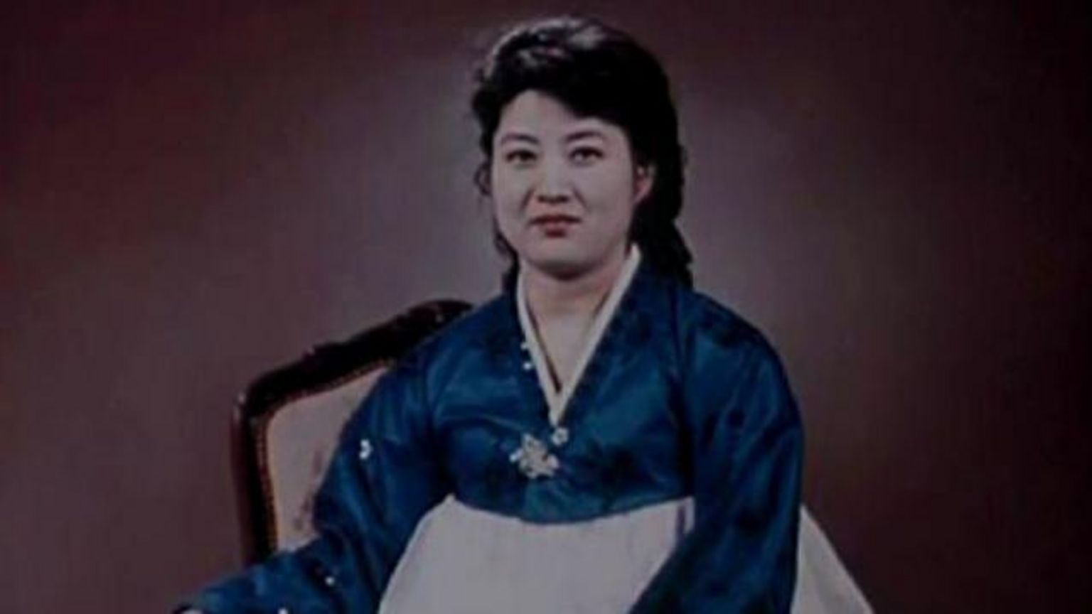 高英姬,大概三、四十岁,身穿蓝白相间的韩服,对着镜头微笑。