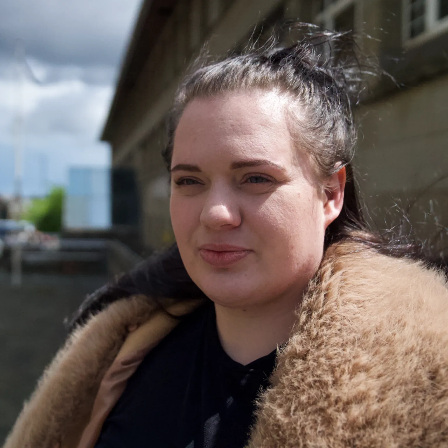 Fiona, a woman with dark hair partly tied back, wearing a jacket with a fur collar - she is outside with a large building behind her