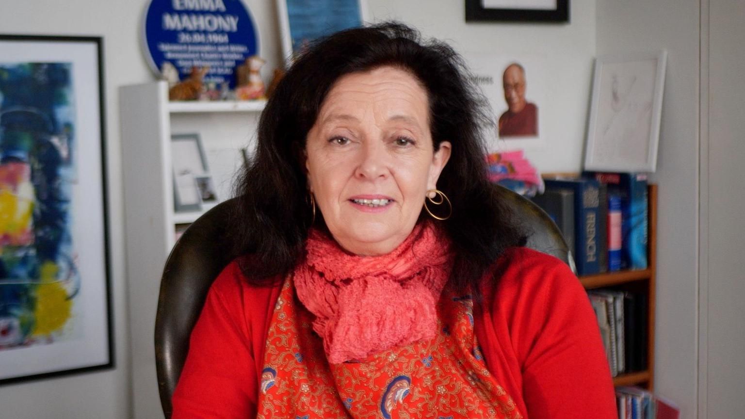 A woman wearing a red shirt and scarf. She has brown hair and is looking at the camera. She is sat in an office in front of a book case and a colourful painting to the left of the image. The walls of the office are white