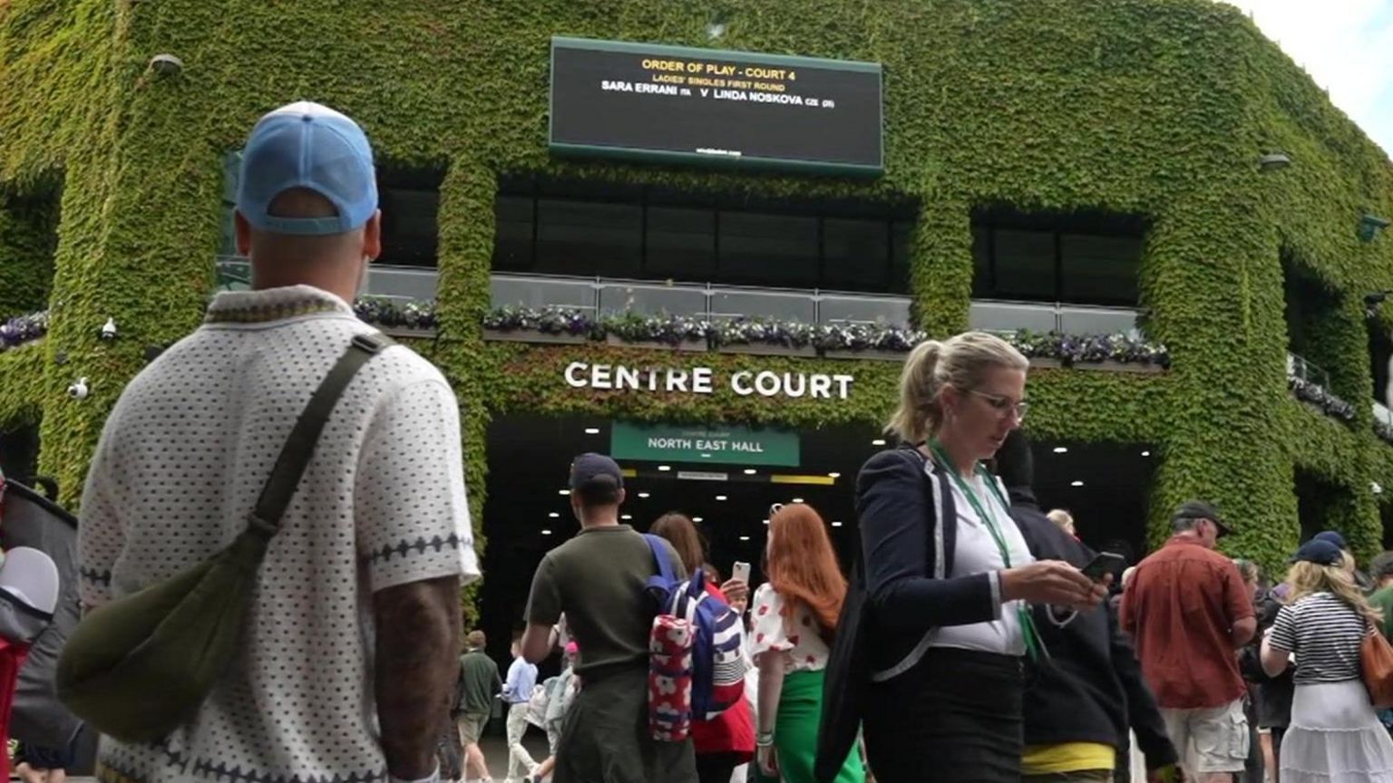 a building covered with green foliage, with 'centre court' written in white writing. A crowd is walking past.