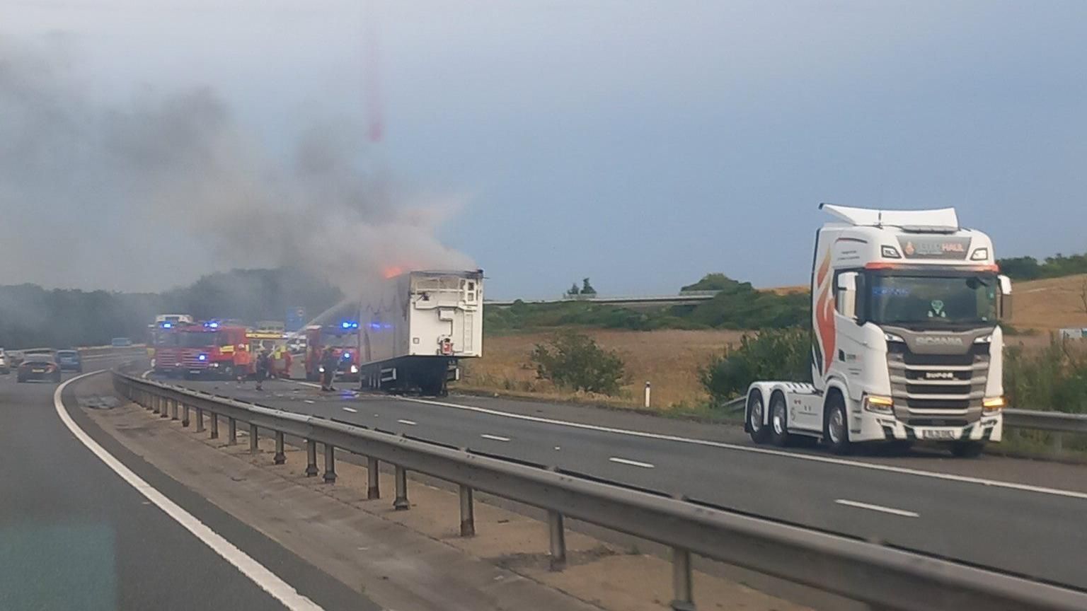 Flames and smoke coming out of a stationary white lorry, as fire engines are parked beside it and firefighters pour water on it out of a hose. 