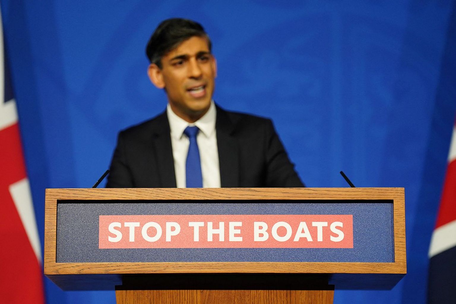 Rishi Sunak standing behind a podium with the slogan STOP THE BOATS emblazoned on it. There are Union flags on either side of him and a blue background.