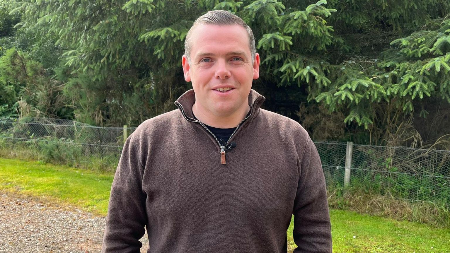 Man in a brown jumper, smiling with both hands in his pockets, standing in a rural-looking area with large trees behind him