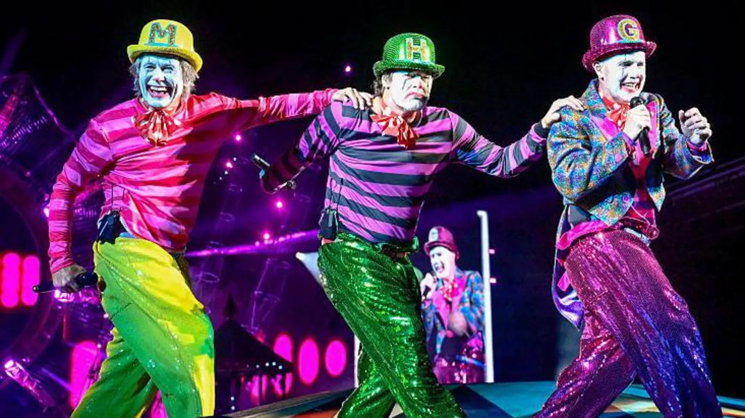 Getty Images The three men in pink and green circus outfits and face paint performing on stage in a row. They are all wearing bowler hats with the first letter of their name on. Gary Barlow holds a microphone up to his mouth, while Mark has his hand on Howard's shoulder, and Howard has his hand on Gary's shoulder. They all wear sequin trousers.