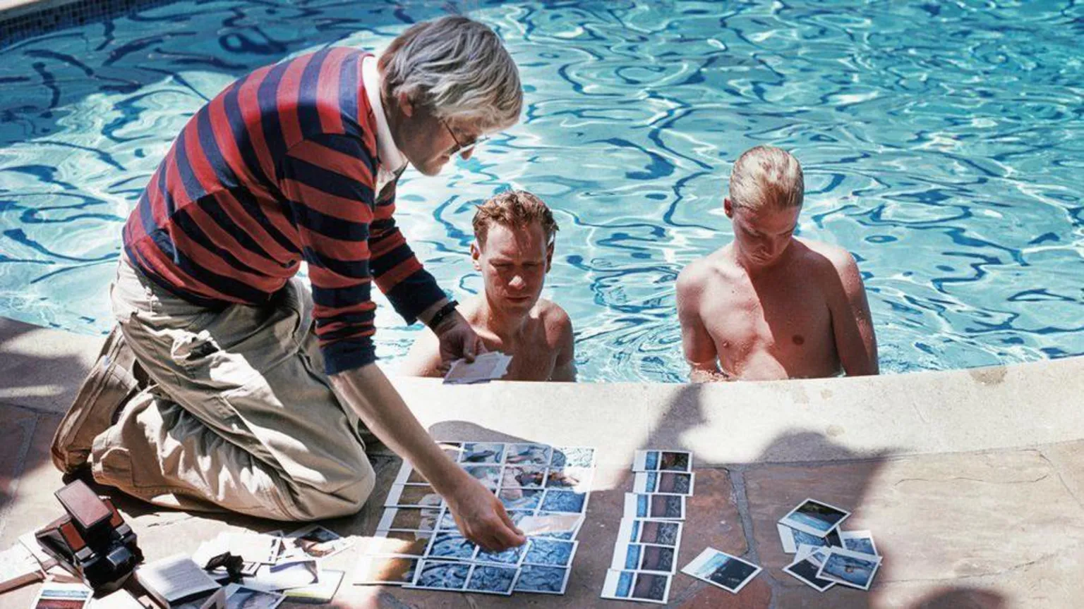 Getty Images David Hockney is shown kneeling by a swimming pool in camera. Two swimmers are watching him piece together photos of the pool.