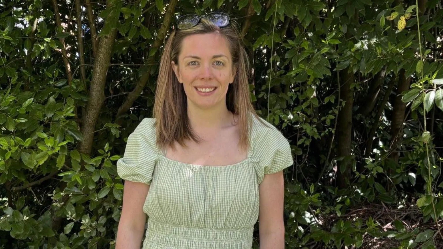 Other A woman with light brown hair wears a green gingham dress with puff sleeves smiles at the camera. She has sunglasses perched on the top of her head.