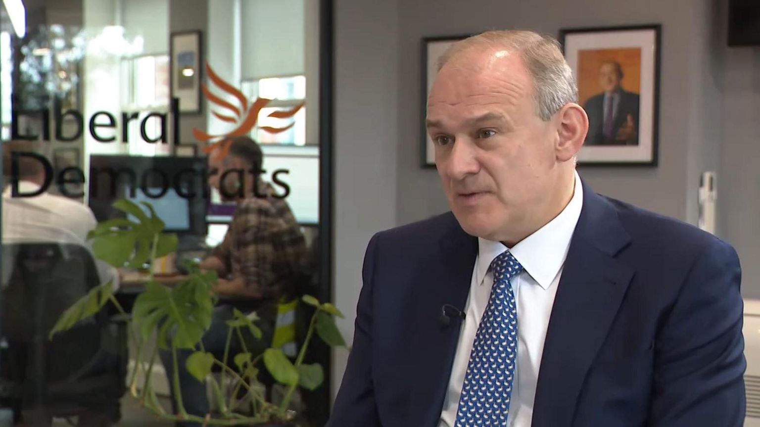 Sir Ed Davey wears a white tie, blue and white shirt, and navy blue suit with a clip-on microphone on the lapel. He is sitting in an office with grey walls and two people working at a desk can been seen through a large glass window on the left. On the glass are the words "Liberal Democrats" in black writing and the party's logo