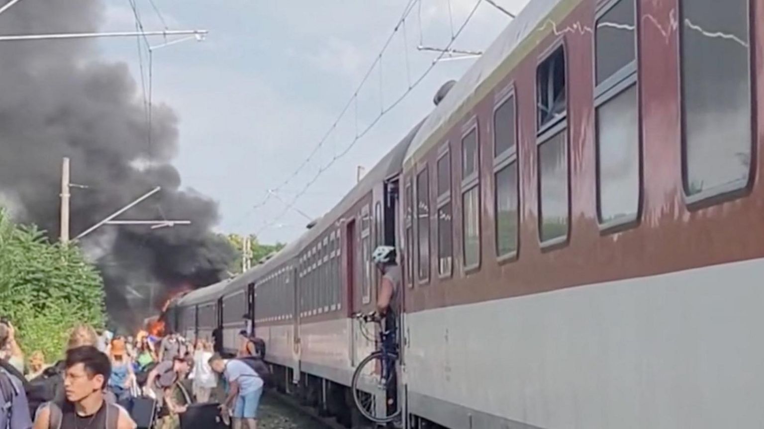 Passengers walk away from a train that is on fire at one end, with one man leaving the train with his bicycle.