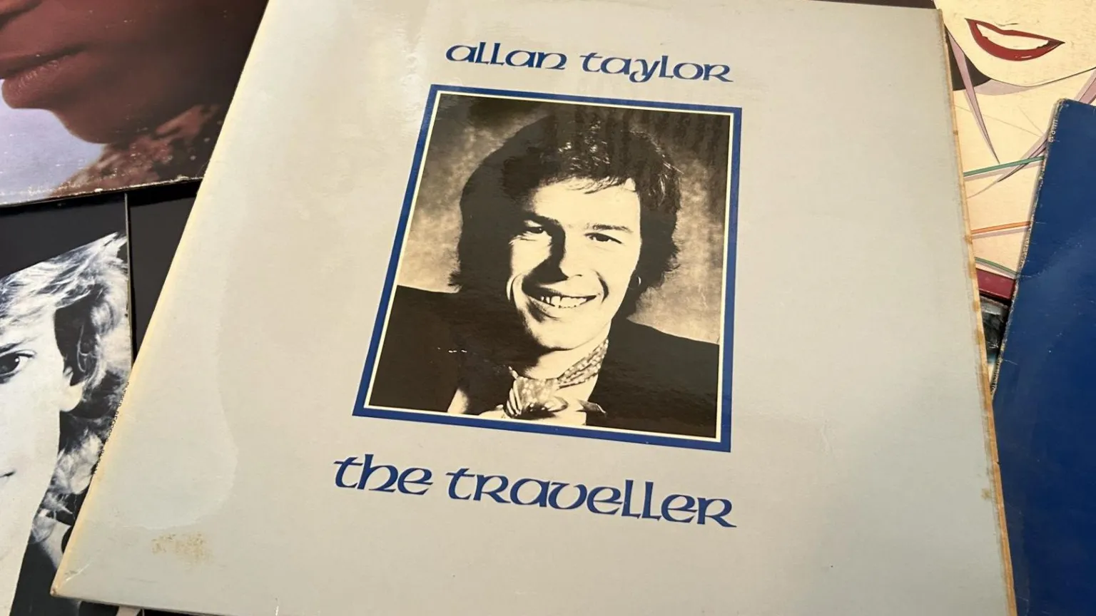 Allan Taylor's LP The Traveller showing a blue cover with a black & white photo of him smiling wearing a neckercheif in a frame in the centre