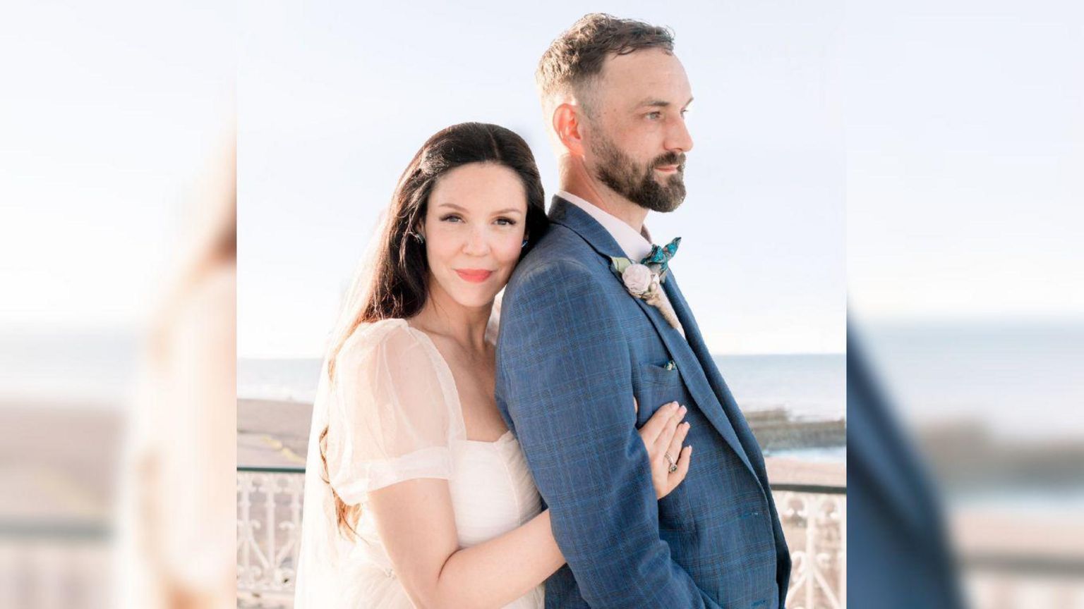 Ally standing behind Paul wearing her wedding dress. She is hugging him from behind and smiling while Paul looks off to the right.