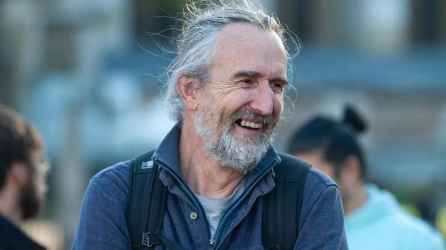 The image shows an older man with gray hair and a gray beard, smiling warmly. He's wearing a dark blue button-up shirt or jacket over what appears to be a gray t-shirt. The background is blurred, showing other people in an outdoor setting.