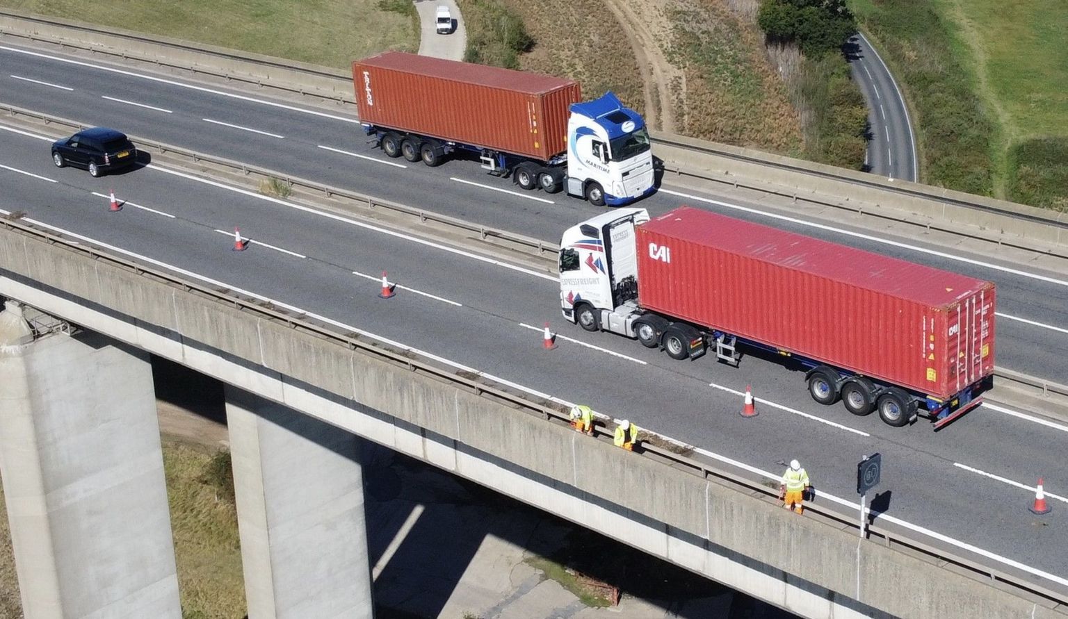 Red lorries travelling in opposite directions on the bridge. One lane is closed on both side and is sectioned off with cones