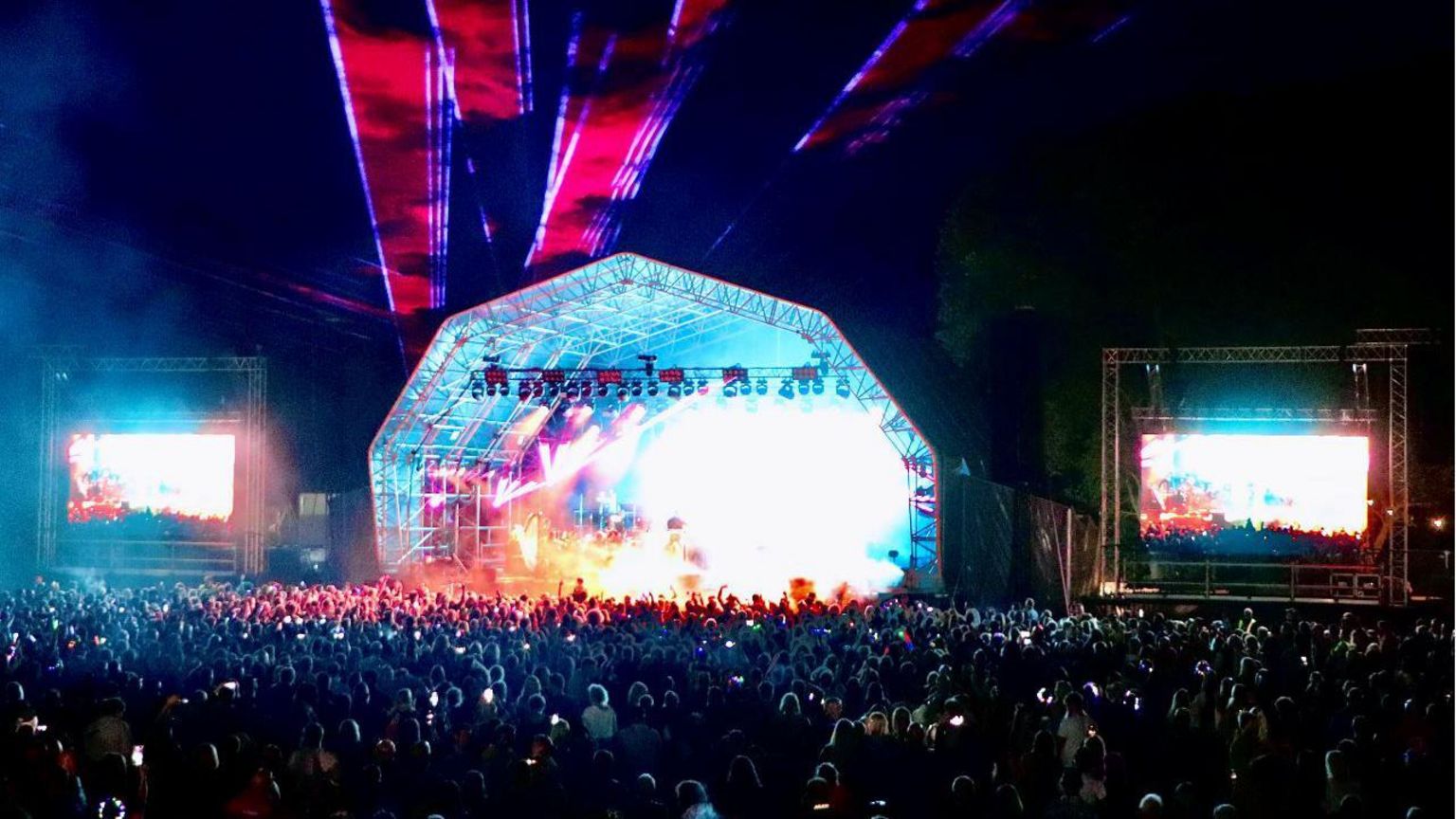 A shot of the main stage at night. There are hundreds of people gathered to watch a pink, white and blue laser light show with smoke machines 