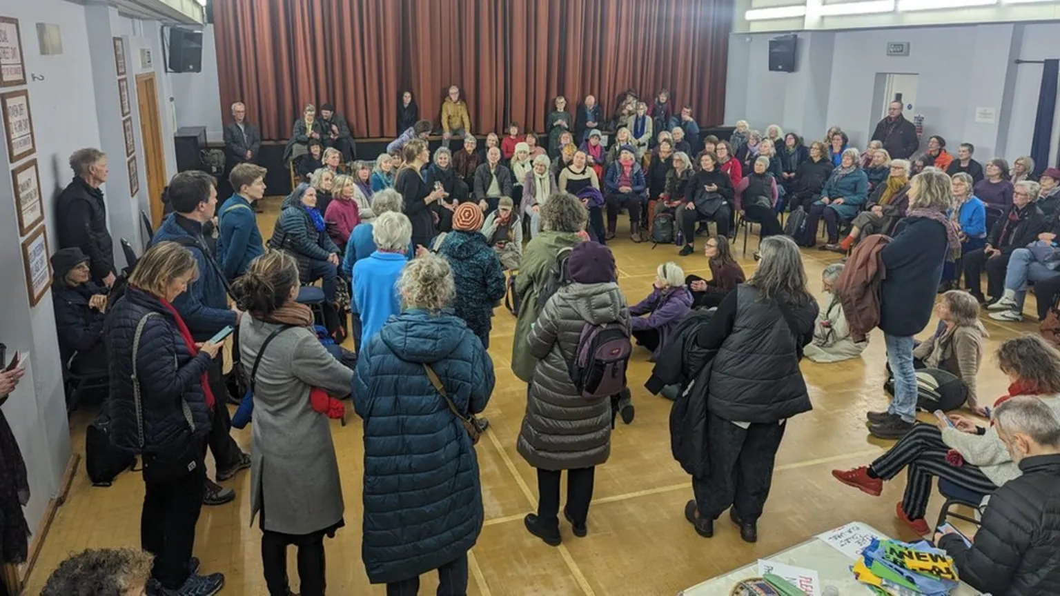 A large group of people gathered in a community hall with wooden floors and red curtains. Many wear winter coats and are arranged in a circle formation around the room's perimeter. Some sit on chairs while others stand, appearing to be participating in a group rehearsal session.