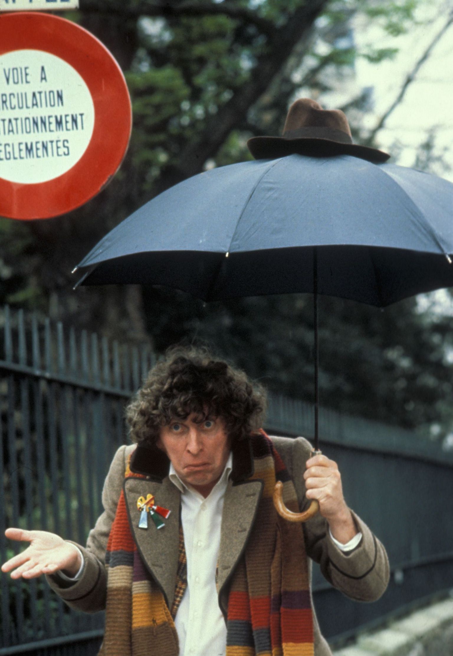 Tom Baker in costume as the 4th Doctor, shrugging his shoulders and carrying an umbrella with the Doctor's hat on top