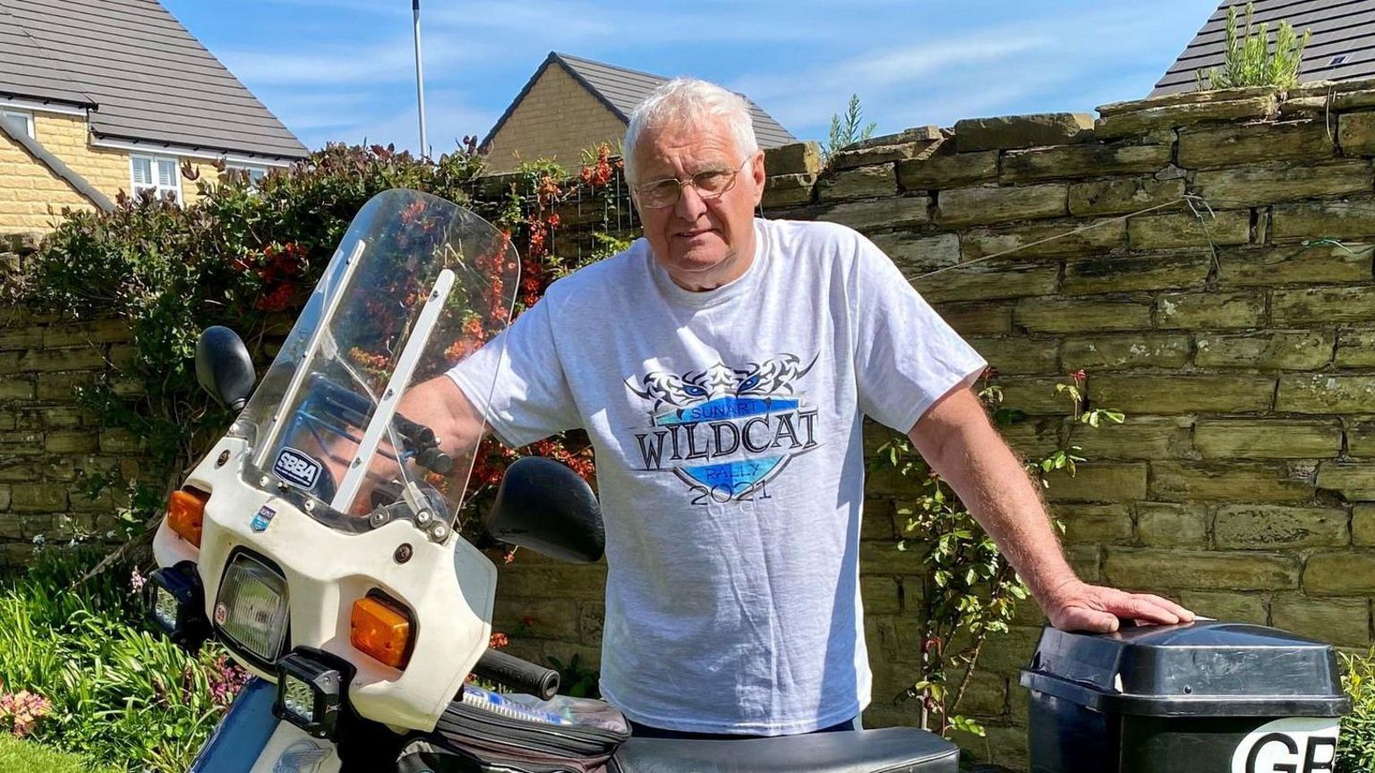 Mr Stanley stand in front of a motorbike. He has short white hair and is wearing glasses