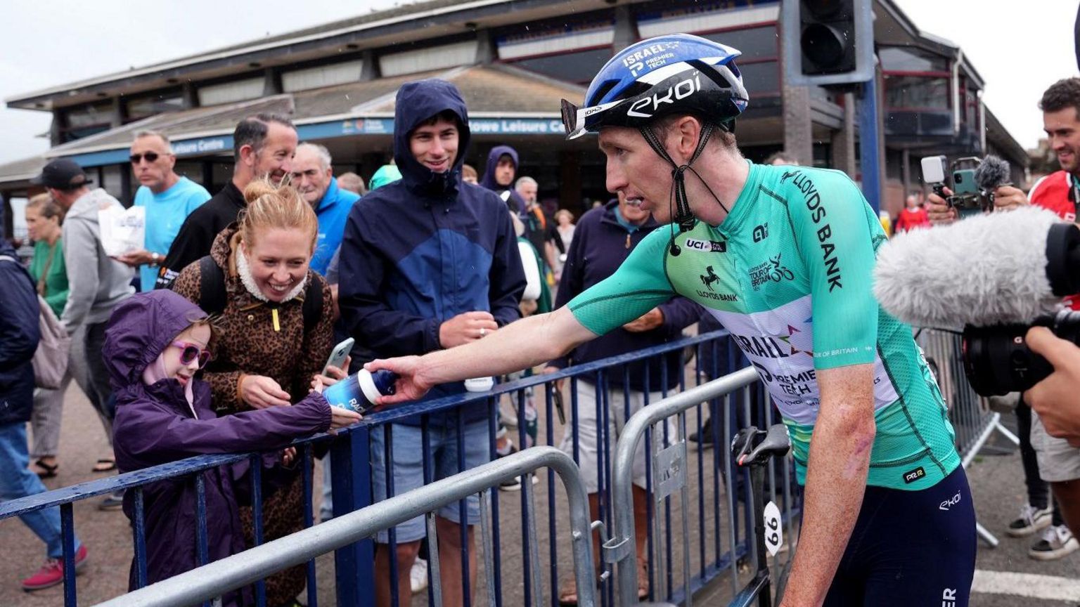 Cyclist gives a water bottle to a girl wearing purple coat and sunglasses while onlookers smile