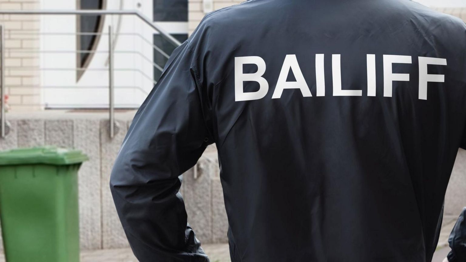 Rear View Of A Male Bailiff Standing With His Hands On Hips At House Entrance