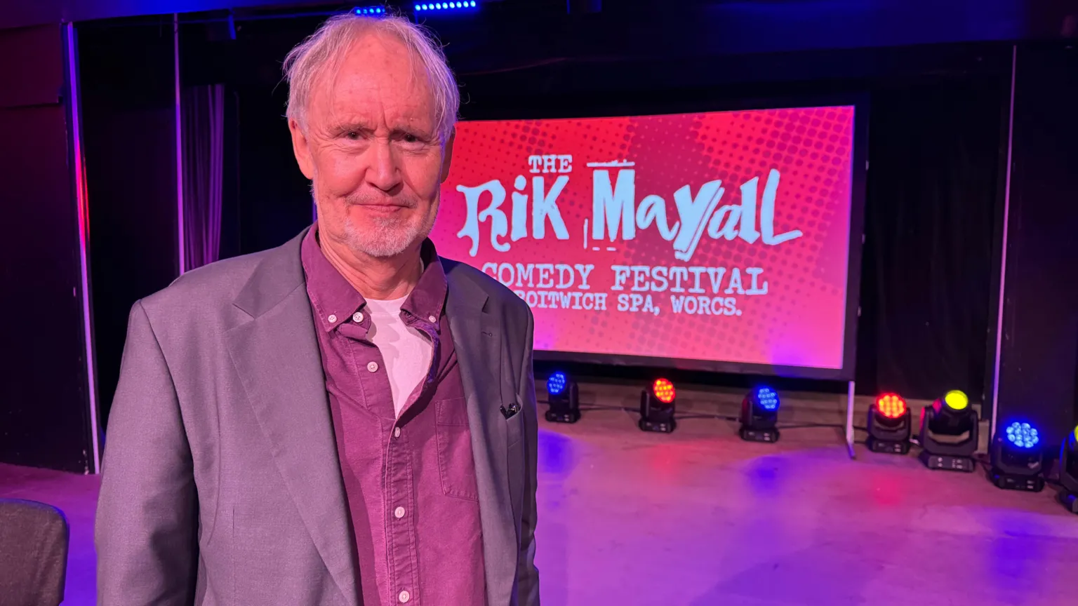 Nigel Planer - an elderly man with short white hair, grey jacket, red shirt over a white t-shirt, stands on a stage and smiles. A screen which says The Rik Mayall Comedy Festival, can be seen behind him.