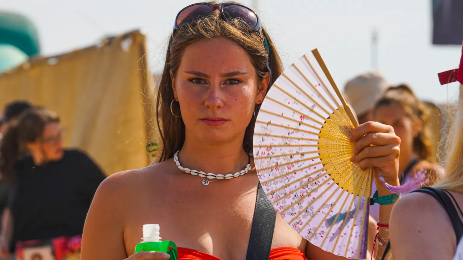 Hugh R Hastings/Getty Images A woman with brown hair stares directly at the camera wearing a red bandeau, using a pink and yellow fan, with sunglasses on and a white shell necklace. In the background other festival goers are blurred