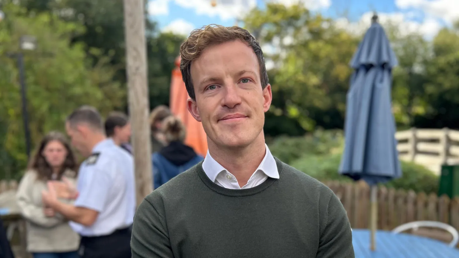 A man wearing a jumper with a white collar shirt underneath. He is looking at the camera and is smiling. He is standing outside