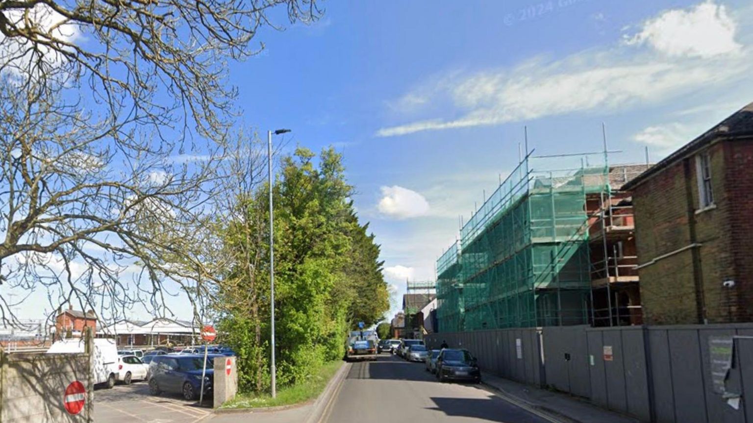 Churchfields Road in Salisbury. A no entry sign by the train station can be seen to the left, whilst buildings in scaffolding can be seen on the right. Cars are parked up on the kerb on the right. 