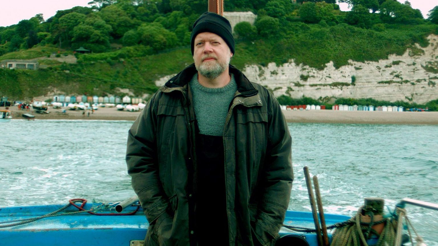 Photo of Paul Breen in character as a fisherman on a boat with the sea and a beach in the background.
