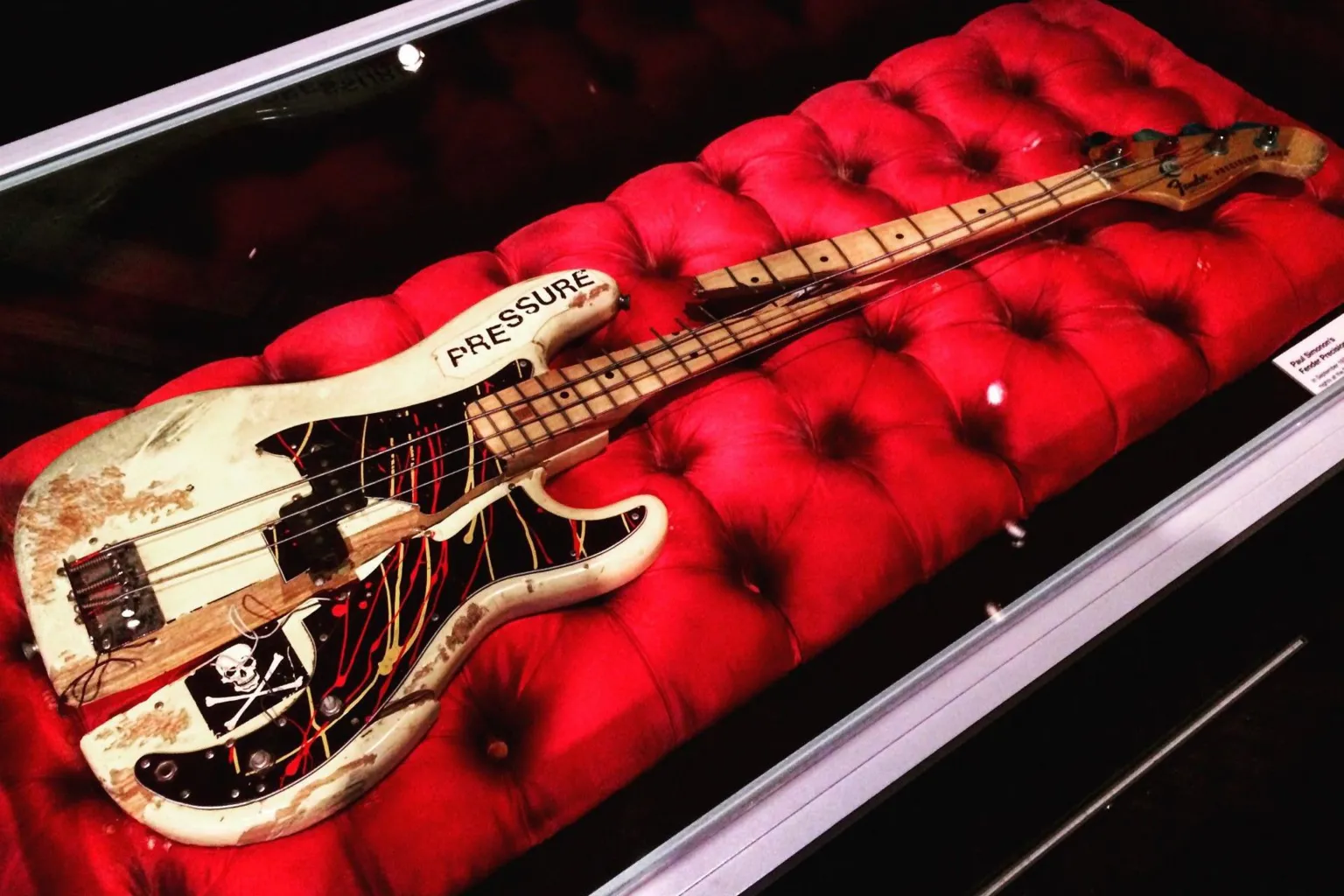 White bass guitar split in two with a skull and crossbones and pressure stickers stuck on it. It is presented on a large red cushion in a glass case