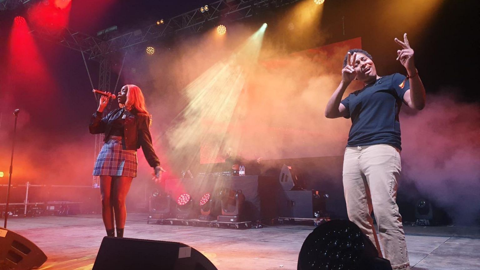 Artist Enny on the left performing on stage, she is holding a mic and is wearing a tartan skirt and a black leather jacket, Ali is on the left and is wearing brown trousers and a black t-shirt, she is using her hands to sign