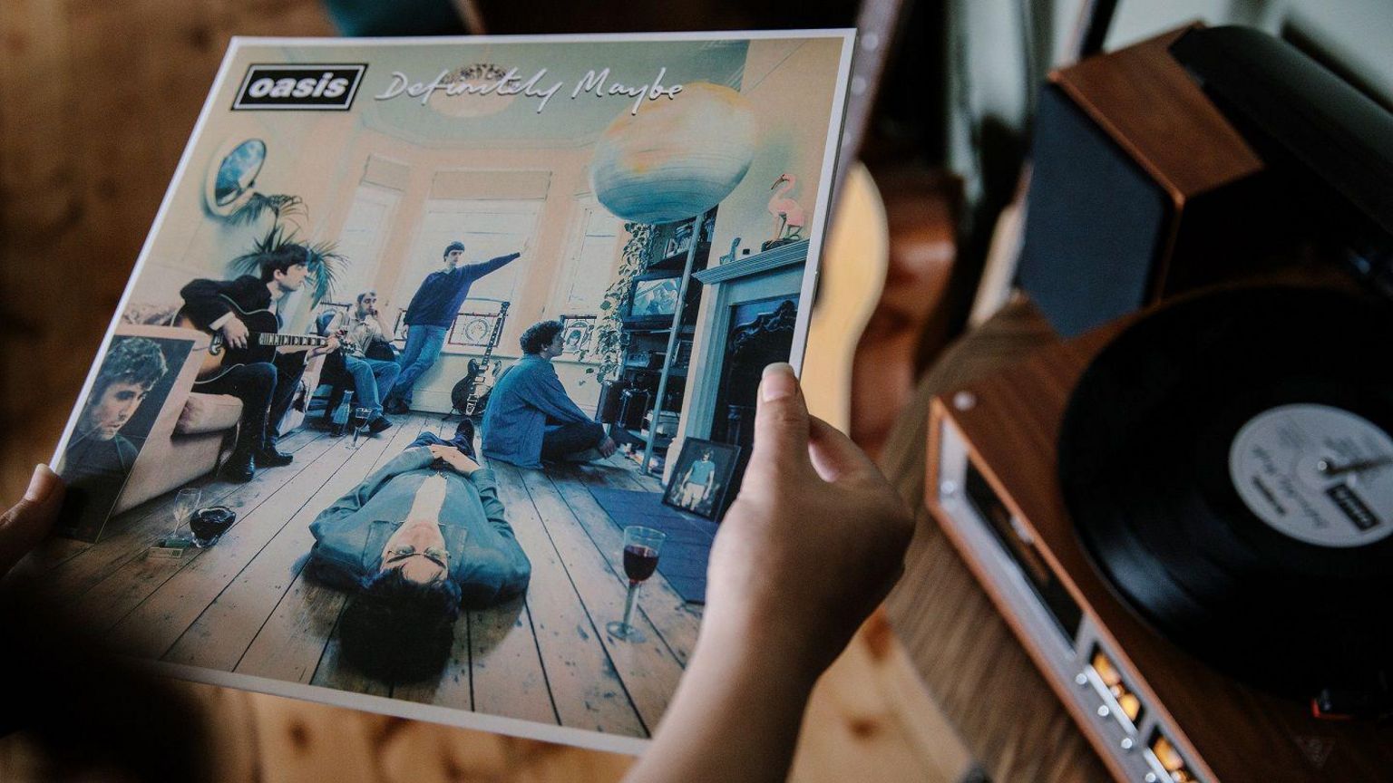 A pair of hands holding Oasis' Definitely Maybe album cover as the vinyl record plays on a nearby turntable. The cover shows Liam Gallagher lying on the wooden floor of a living room with a glass of what appears to be red wine next to him. Noel Gallagher is sat on a couch holding a guitar. A picture of Burt Bacharach is next to the couch. Three other members of the band are in various poses, one sat on the floor looking at a TV, one sat on a chair and another standing in front of the window.