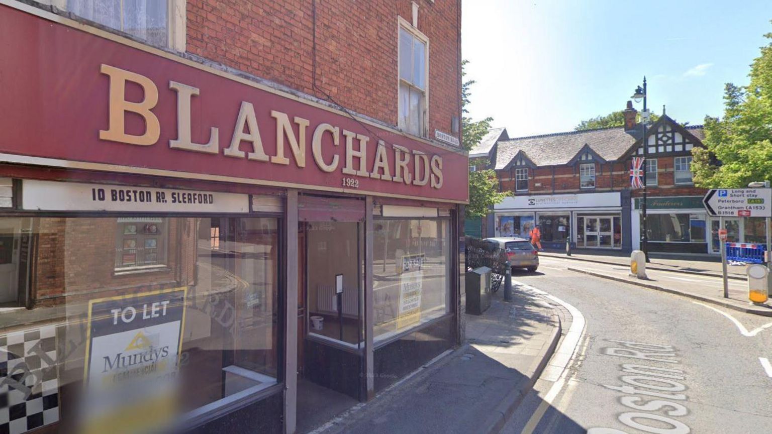 The front of Blanchards coffee shop, with Blanchards written in gold capital letters and on a dark red background. There is a To Let sign in the window and the shop is situated on a bend with a road sign opposite pointing left towards Grantham and the train station