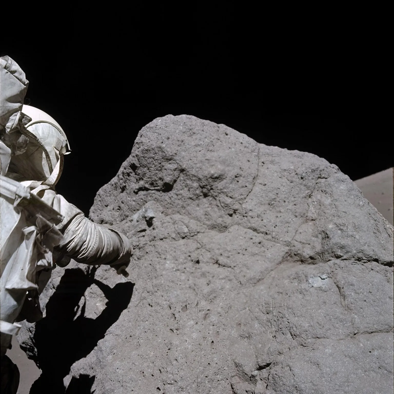 NASA The Apollo astronaut Harrison Schmitt, from Apollo 17,  is collecting samples  rock on the Moon. The rock is slightly taller than him and about 3 times as wide. He is on the left of the photo with his back to the camera, and is wearing a white NASA space suit as he tries to collect the sample using his right arm. His body is creating a shadow on the large dark grey rock. At the top of the photo you can see the blackness of space.